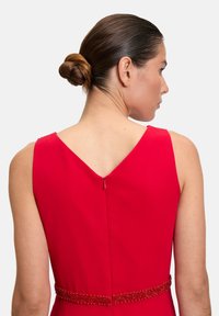 Rotes Kleid mit V-Rücken-Design, ausgestattet mit einem versteckten Reißverschluss und einem mit Perlen besetzten Taillenbund für zusätzliche Details. Glatte Stofftextur.