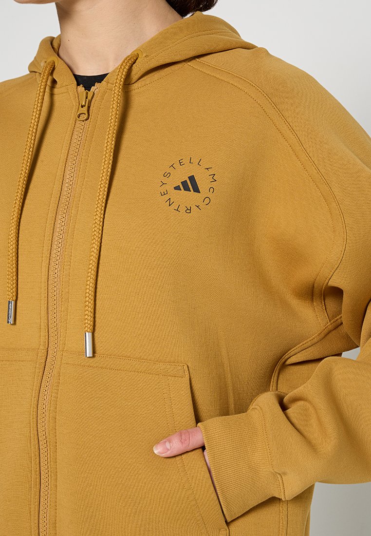 Sudadera con cremallera de color mostaza, con un bolsillo frontal, fabricada con un tejido texturizado y un logotipo negro circular en el lado izquierdo. Presenta cordones trenzados.