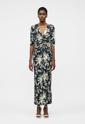 Γυναίκα που φοράει μεσοφόρι με μπλε ναυτικό χρώμα και φλοράλ σχέδιο, με ζαρτιέρες στρόγγυλου στρέτσινγκ στο μπροστινό μέρος, ντεκολτέ σε σχήμα V, μανίκια μέχρι τους αγκώνες και λευκά πέδιλα με λουριά στον αστράγαλο και ανοιχτά δάχτυλα.