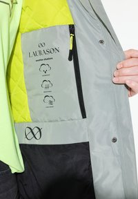 Grijze gewatteerde interieur met een felgele paneel. Bevat ritsen, iconen voor waterdichte, ademende en winddichte eigenschappen. Logo zichtbaar.