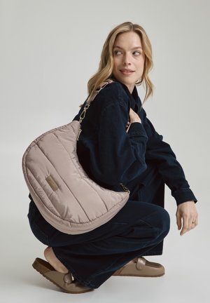 Sac en bandoulière en rose clair matelassé, de forme croissant, avec des accessoires dorés et une bandoulière amovible, associé à une tenue en denim foncé.
