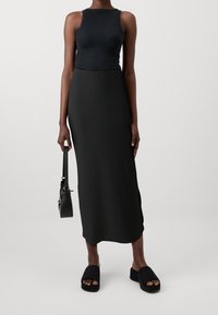 Schwarzes geripptes Tanktop, langer schwarzer gerippter Rock, schwarze Rutschschuhe und eine kleine schwarze Handtasche. Einfaches, figurbetontes Design mit strukturiertem Finish.