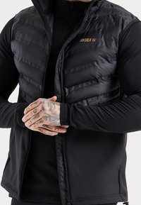 Černá bezrukávová péřovka s horizontálním prošíváním, zapínáním na zip a zlatým logem. Nositá na fitted černém dlouhém rukávu.