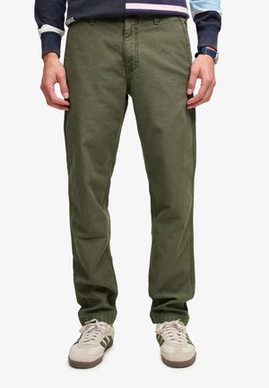 Homme portant un pantalon décontracté vert olive et des baskets blanches avec des bandes vertes, debout sur un fond blanc.