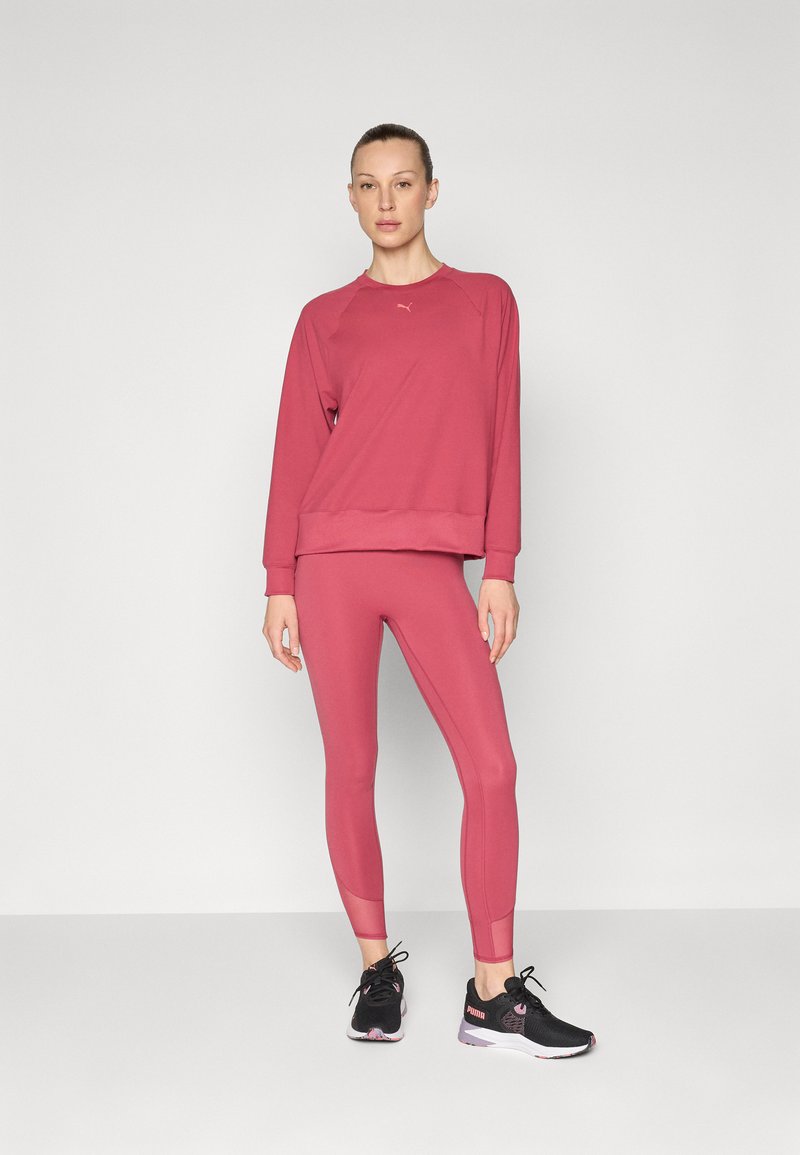 Frau in einem rosa Sweatshirt und passenden Leggings, beide aus einem glatten, dehnbaren Material. Schwarze Sportschuhe runden das Outfit ab.