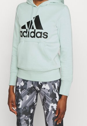 Persoon met een lichtgroene Adidas-hoodie met voorzak en zwart-wit-grijs abstract patroon op de legging, getoond van schouders tot dijen.