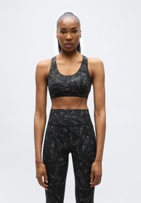 Αθλητική γυναίκα με μαλλιά μαζεμένα πίσω, φοράει αθλητικό σουτιέν με μαύρο και γκρι μοτίβο και κολάν με ψηλή μέση, στέκεται μπροστά σε ένα απλό φόντο.