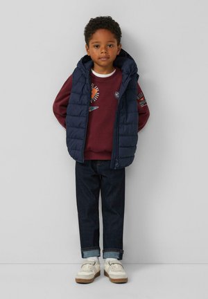 Marine blauwe puffer vest over een bordeaux sweatshirt, donkere spijkerbroek en witte sneakers. De vest heeft een capuchon; het sweatshirt heeft geprinte graphics.