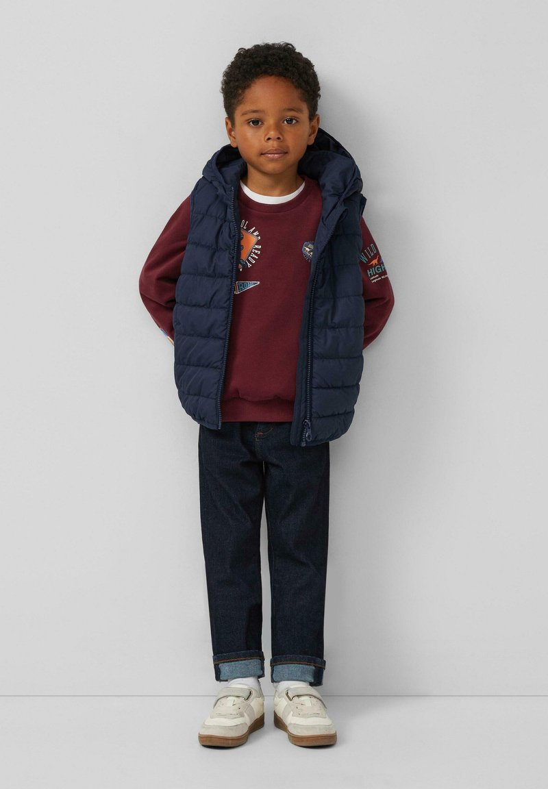 Gilet imbottito navy su felpa bordeaux, jeans in denim scuro e sneaker bianche. Il gilet è dotato di cappuccio; la felpa presenta grafiche stampate.
