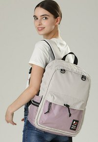 Zaino grigio chiaro con una tasca inferiore viola, cerniere nere e spallacci. Presenta una toppa con logo nera e chiusura a cordoncino sulla tasca anteriore.