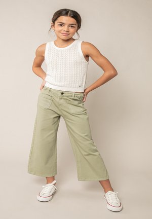 Jeune fille debout, mains sur les hanches, portant un haut en tricot sans manches blanc, un pantalon large vert clair et des baskets blanches.