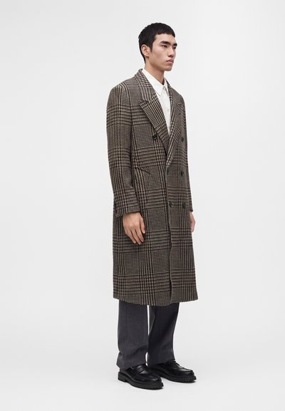 Homme debout portant un long manteau marron et noir à carreaux, une chemise blanche, un pantalon gris et des chaussures noires sur un fond clair uni.