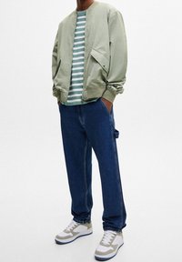 Giacca bomber verde sopra una maglietta a righe, abbinata a pantaloni in denim blu e sneakers di colore chiaro. Texture lisce e design casual.