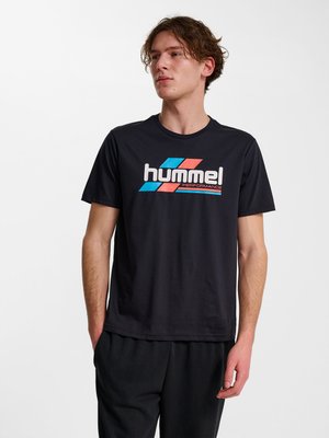 Ung mand med lys hud og bølget hår iført en sort t-shirt med blå og rød "hummel performance" logo, stående med den ene hånd i lommen.