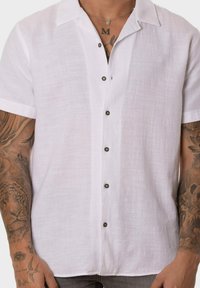 Camisa de lino blanca de manga corta con cierre de botones en la parte delantera, que presenta botones oscuros y una tela suave y texturizada.