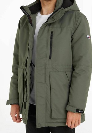 Homme portant une veste d'hiver ouverte de couleur olive avec fermeture éclair et poches, par-dessus un t-shirt blanc et un pantalon noir.