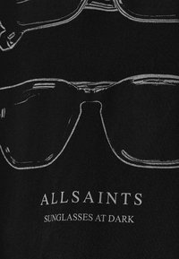 Schwarzer Stoff mit weißen Umrissen von Sonnenbrillen, dazu der Text "ALLSAINTS" und "SONNENBRILLEN IM DUNKELN" unter dem Design.