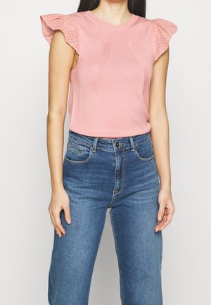 Femme portant un haut en maille rose sans manches avec des manches à jour en volants et un jean bleu taille haute, debout devant un fond uni.