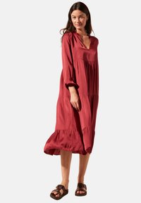 Robe fluide rouge avec un décolleté en V, de longues manches bouffantes et des ruffles en couches à l'ourlet. Le tissu semble doux avec un léger brillant. Sandales marron.