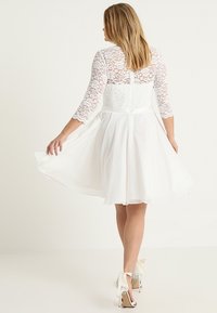Bílé šaty s krajkovými 3/4 rukávy, fitted korzetem, rozšířenou sukní a průhledným krajkovým zadním dílem. Spárovány s bílými vysokými podpatky s mašlemi.