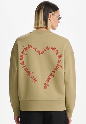 Person mit kurzen schwarzen und blonden Haaren trägt einen beigefarbenen Pullover mit rotem, herzförmigem Text auf dem Rücken, der von Wellen und Meer handelt.