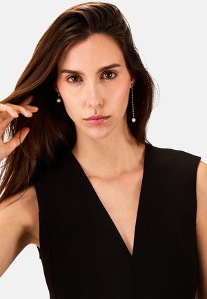 Sort ærmeløs kjole med V-hals, struktureret stof og synlig syning. Modellen bærer perle-drops øreringe, har langt brunt hår og neutral makeup.