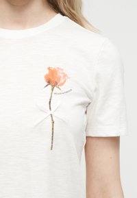 Λευκή μπλούζα με τύπωμα πορτοκαλί τριαντάφυλλου, μικρό λευκό φιόγκο στο στελέχος, και κείμενο "PÉTALES & POSITIVITÉ" στην περιοχή του στήθους.