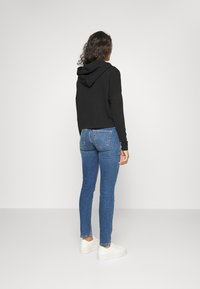 Kvinna med lockigt mörkt hår, iförd en svart hoodie, blå tajta jeans och vita sneakers, stående vänd mot en tom vit vägg.