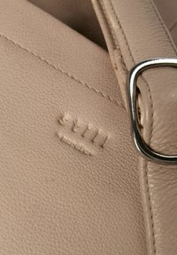 Details van een beige leren handtas met een gladde textuur, opvallende stiksels en metallic gespsluiting. De merknaam is in reliëf aangebracht.