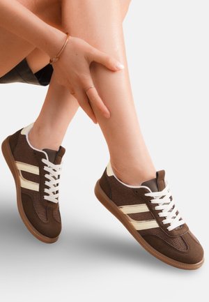Personne portant des baskets marron et beige avec des lacets blancs, touchant leur bas de jambe, sur un fond clair uni.