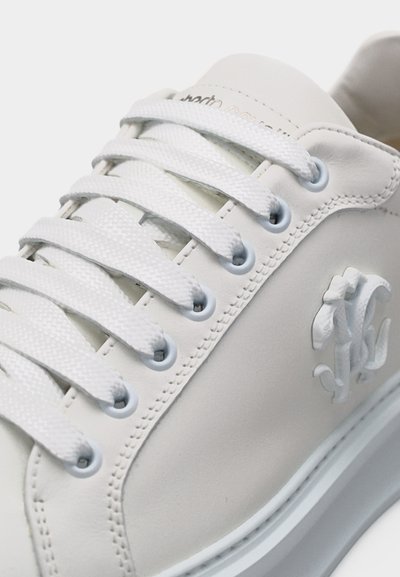 Κοντινό πλάνο λευκού δερμάτινου αθλητικού παπουτσιού με λευκά κορδόνια, μεταλλικά φερμουάρ, ραμμένες λεπτομέρειες και ανάγλυφο λογότυπο της μάρκας στο πλάι.