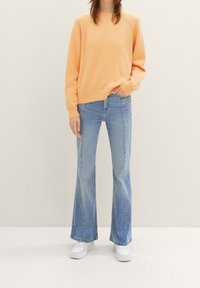 Pomarańczowy sweter o fakturze, jasnoniebieskie rozszerzane dżinsy i białe trampki. Strój charakteryzuje się luźnym krojem i prostymi, czystymi liniami.