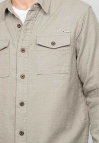 Chemise boutonnée beige clair en tissu texturé, dotée de deux poches poitrine, de boutons marron et d'un petit accent métallique.
