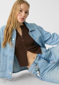 Veste en denim bleu clair par-dessus un top côtelé marron avec des boutons, assortie à un pantalon en denim bleu clair. Détails remarquables incluant un porte-clés de dessin animé sur la ceinture.