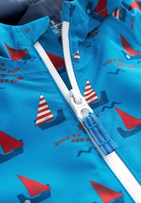 Tissu bleu avec une fermeture éclair blanche, orné d'imprimés de voiliers rouges et blancs et le texte « Il est temps de mettre les voiles » en rouge.