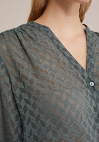 Bluză gri transparentă cu un model geometric, decolteu în V și închidere cu butoane în față. Material texturat cu un luciu subtil.