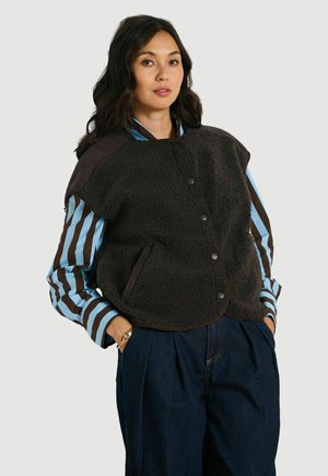 Frau trägt eine schwarze Fleeceweste über einem blau-braunen gestreiften Langarmshirt mit dunkelblauer, hochtaillierter Hose, Hände in den Taschen.