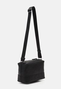 Czarna skórzana torebka w kształcie prostokąta, z metalowymi akcentami w kolorze srebrnym, regulowanym paskiem oraz wytłoczonym napisem "JEANS" wzdłuż dolnej krawędzi.