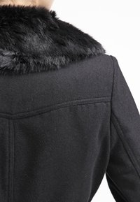 Svart ullblandad kappa med en plysch svart fuskpälskrage, strukturerade axlar och detaljerad bakseam för ett skräddarsytt utseende.