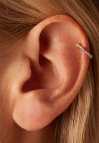 Silberner Creolenohrring mit kleinen, eingelassenen klaren Steinen, aus der Nähe am Rand eines menschlichen Ohrs sichtbar, das die glatte Haut- und Haard textur zeigt.