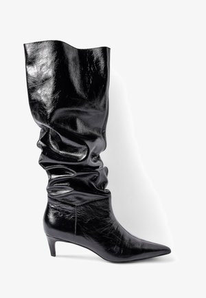 Juodos odinės iki kelių kojinės su laisvu dizainu, smailiu priekiu ir žemu kitten kulnu, demonstruojančios blizgantį, tekstūruotą paviršių.