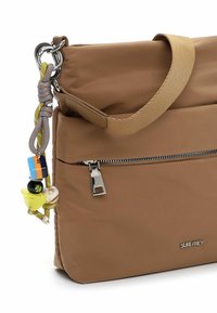Brązowa nylonowa torebka o gładkiej fakturze, z przednią kieszenią zapinaną na suwak, materiałowym paskiem oraz ozdobnym zawieszkiem z kolorowymi koralikami.