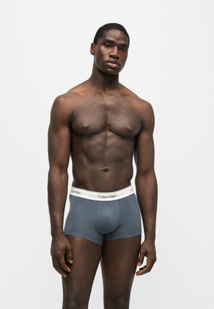 Homme musclé à la peau foncée debout devant un fond uni portant un boxer bleu Calvin Klein avec une ceinture blanche.