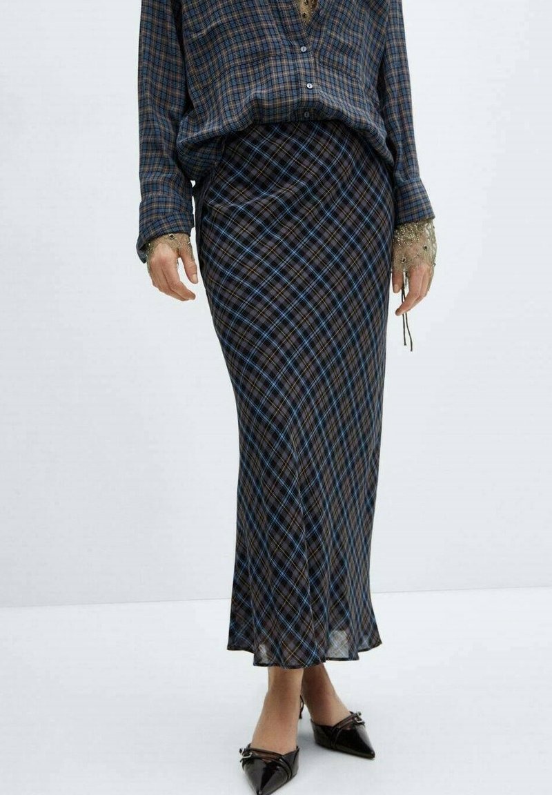 Femme portant une longue jupe à carreaux bleus et bruns avec une chemise ample assortie, des poignets en dentelle et des chaussures slip-on noires à bout pointu.