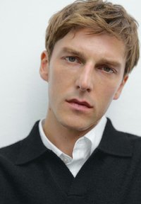 Een close-up van een mannelijke model die een zwart, gebreid sweater met een kraag over een wit shirt draagt, met een zachte textuur en een neutrale achtergrond.