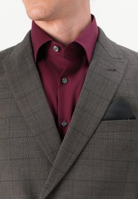 Brown plaid suit jacket with a darker maroon shirt underneath. Features a black pocket square and black buttons on the shirt.