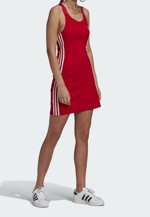Femme portant une robe rouge sans manches avec des bandes blanches sur les côtés et des baskets blanches avec des bandes noires, debout sur un fond clair uni.