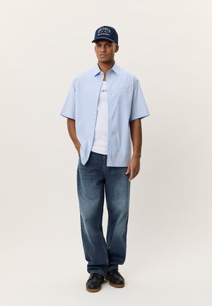 Joven con camisa de manga corta azul claro sobre camiseta blanca, jeans azul oscuro, zapatos negros y gorra de béisbol azul marino, de pie frente a un fondo liso.