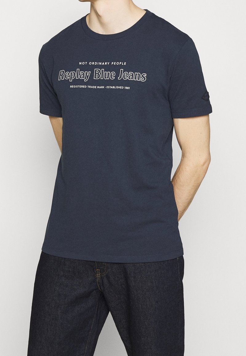 T-shirt en coton bleu marine avec un col rond, arborant un texte blanc "Pas des gens ordinaires, replays en jeans bleus" sur le devant. Manches courtes.