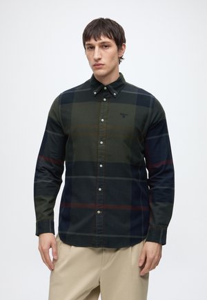 Jeune homme portant une chemise à carreaux boutonnée vert foncé et bleu marine avec un pantalon beige, debout devant un fond blanc uni.